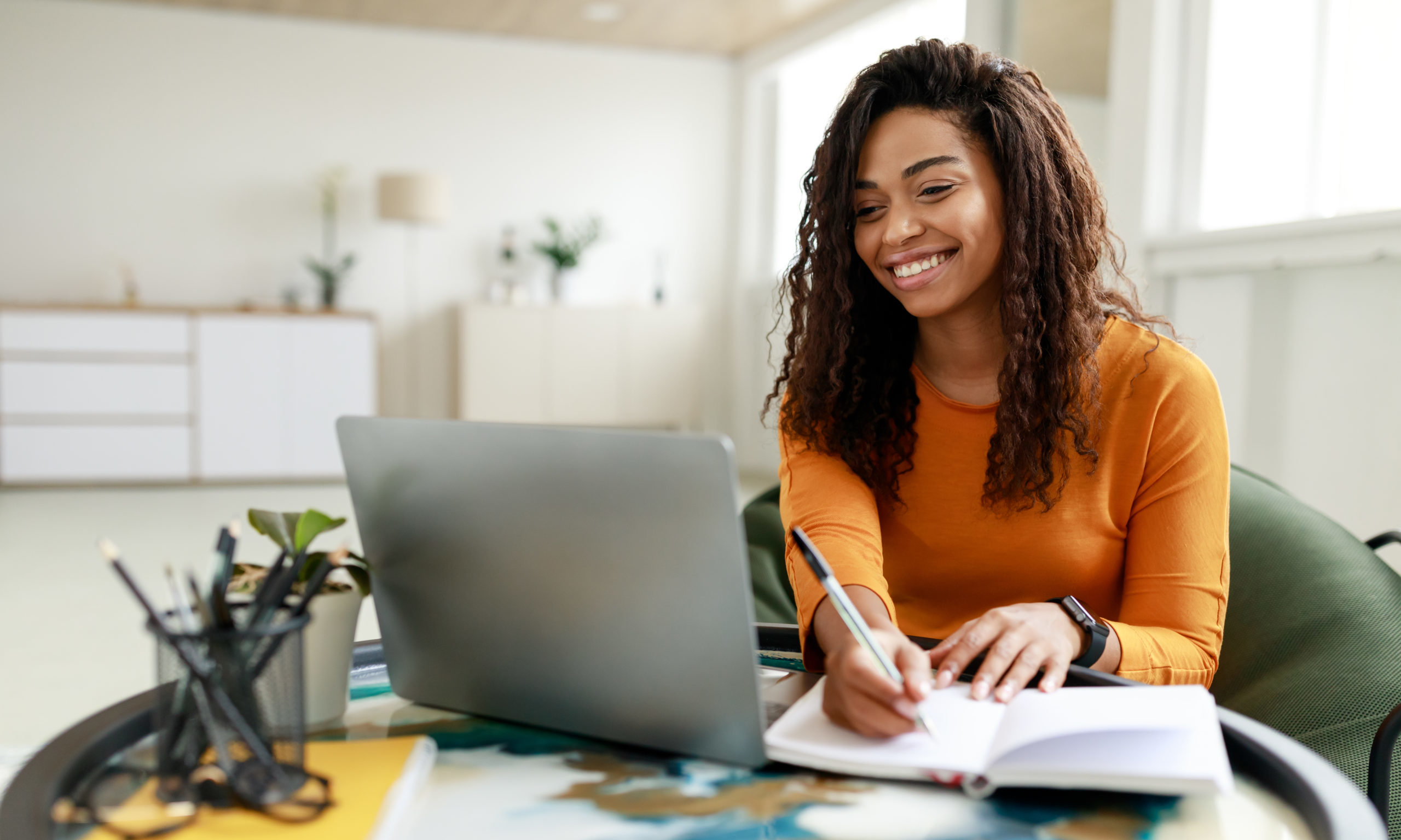 Black woman sitting at desk, using pc writing in notebook Smiling young African American woman sitting at desk working on laptop taking notes in notebook, happy millennial female studying online, watching webinar using computer and writing check list